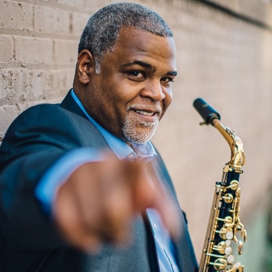 Gray-haired person with a well-groomed beard and a saxophone smiles and points at the camera