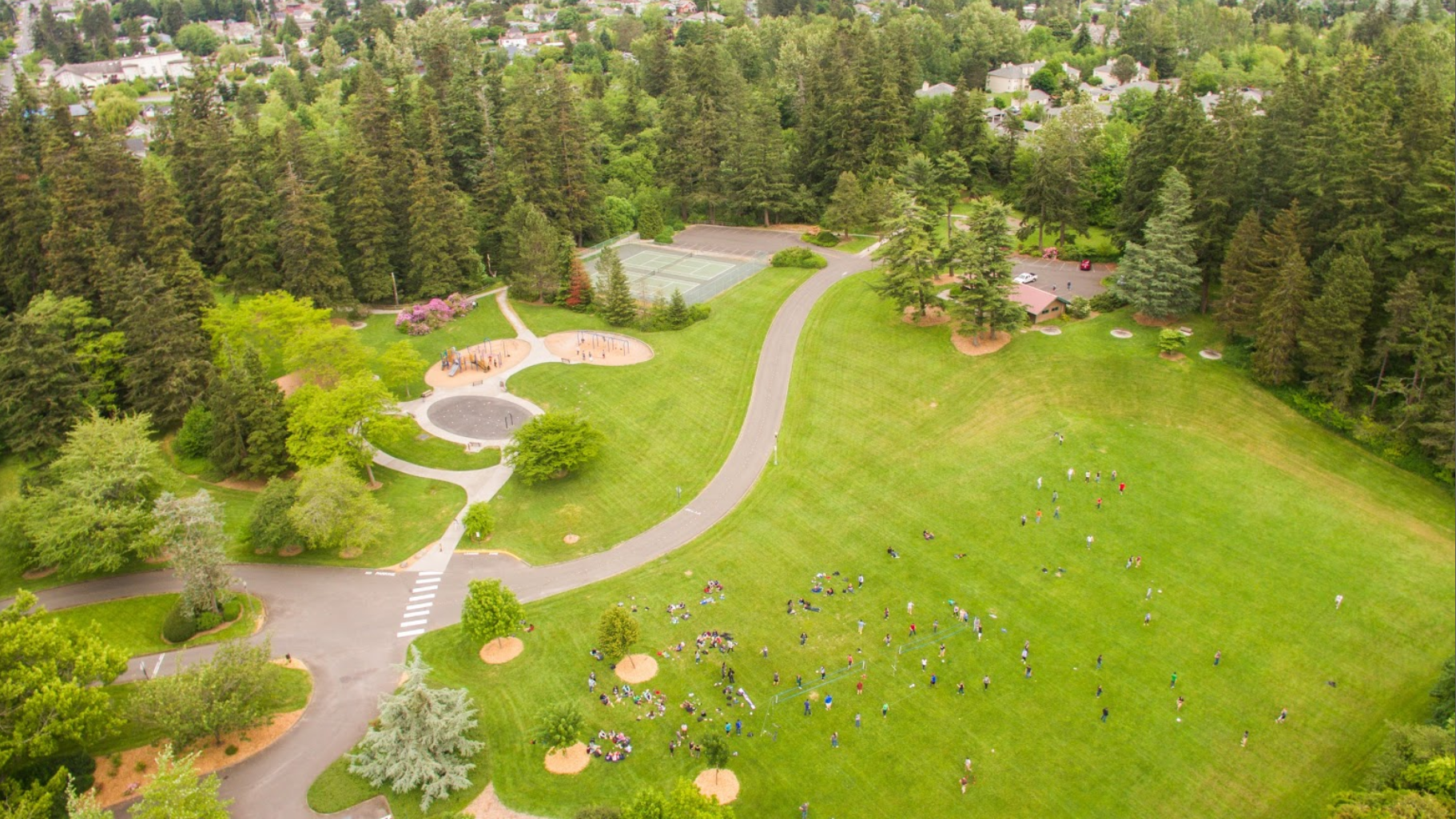 aerial view of Fairhaven Park: a playground and tennis courts on the left, lots of people enjoying a large grassy field on the right, and a shelter with parking lot at the far edge