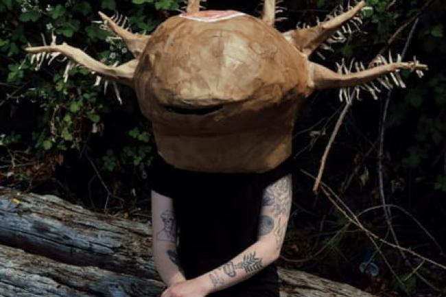 a sculpted fantastical creature head with hairy tentacles, worn as a helmet by a person sitting cross-legged and cross-armed on a log