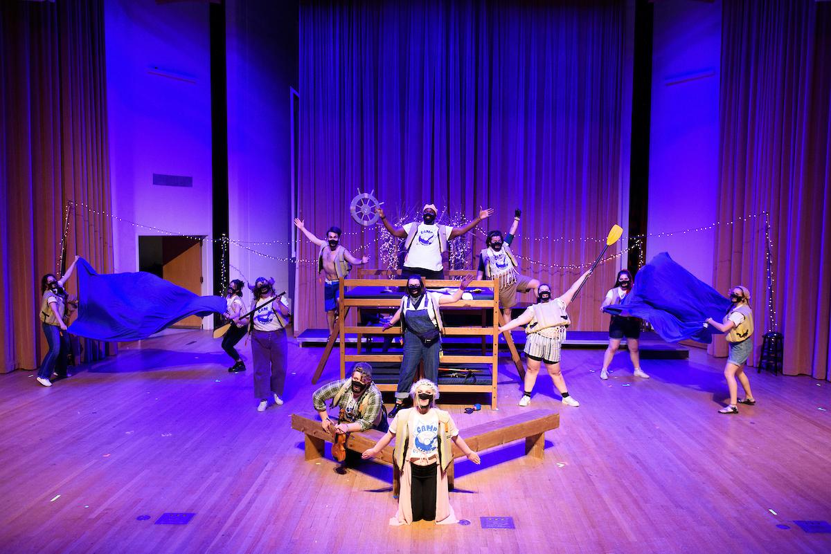Actors posing together on-stage. Stage props include bunk beds, a bench, a boat steering wheel and paddles.