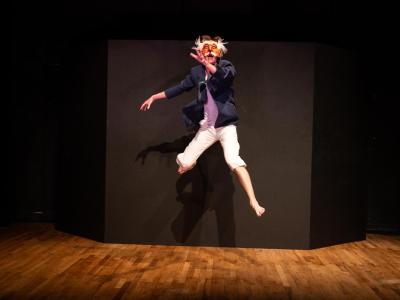 actor in tights, sportcoat and clownish mask leaps into the air
