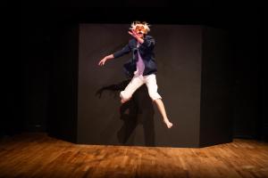 actor in tights, sportcoat and clownish mask leaps into the air