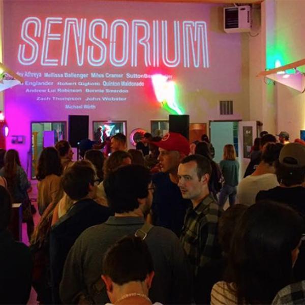neon lit gallery crowded with people. sign on back wall reads "sensorium"