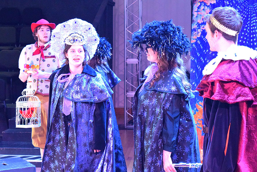 several actors in fanciful red and purple cloaks. Two wear large headresses.