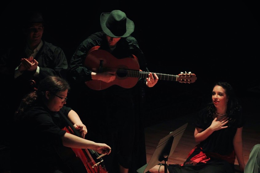 a dark scene of guitarist in hat performing for a seated person.