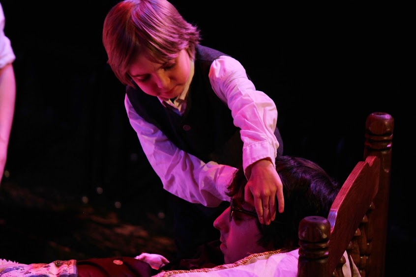 small child holds the head of an actor reclining in bed.