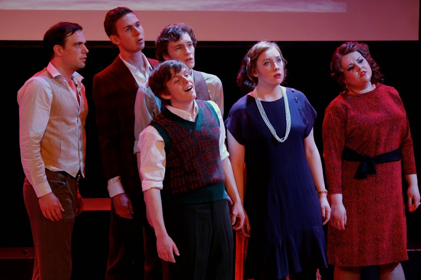 six actors with heads tilted sing together looking out of the frame to the right