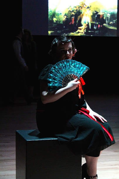 dramtatically lit actor sitting, holding a paper fan
