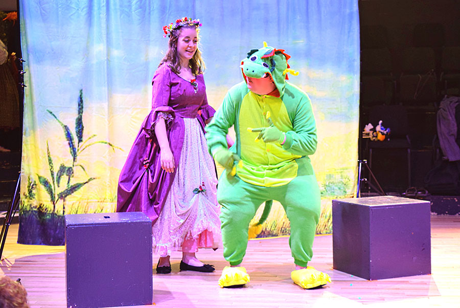 actor in purple dress, actor in dragon suit, two purple boxes against scenic backdrop