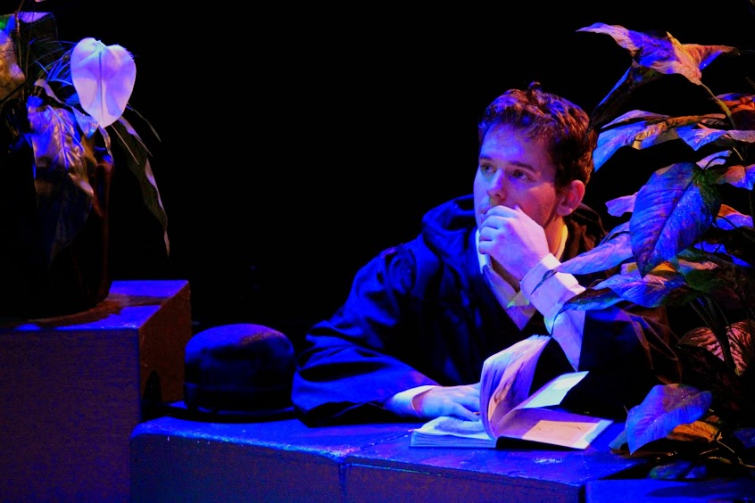contemplative actor seated at desk muses over a book in a deep blue light.