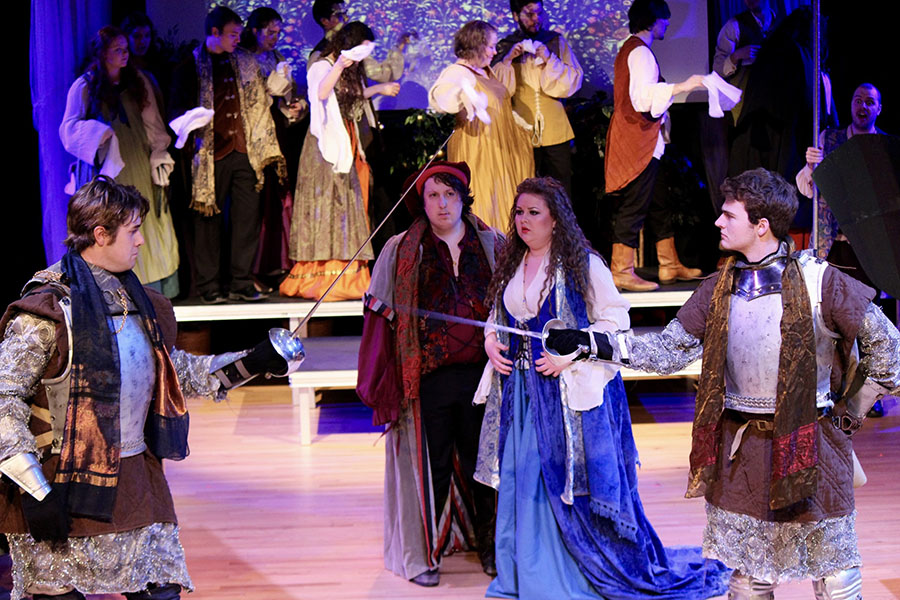 two actors cross fencing swords in front of crowd scene with opera singers in renaissance costume