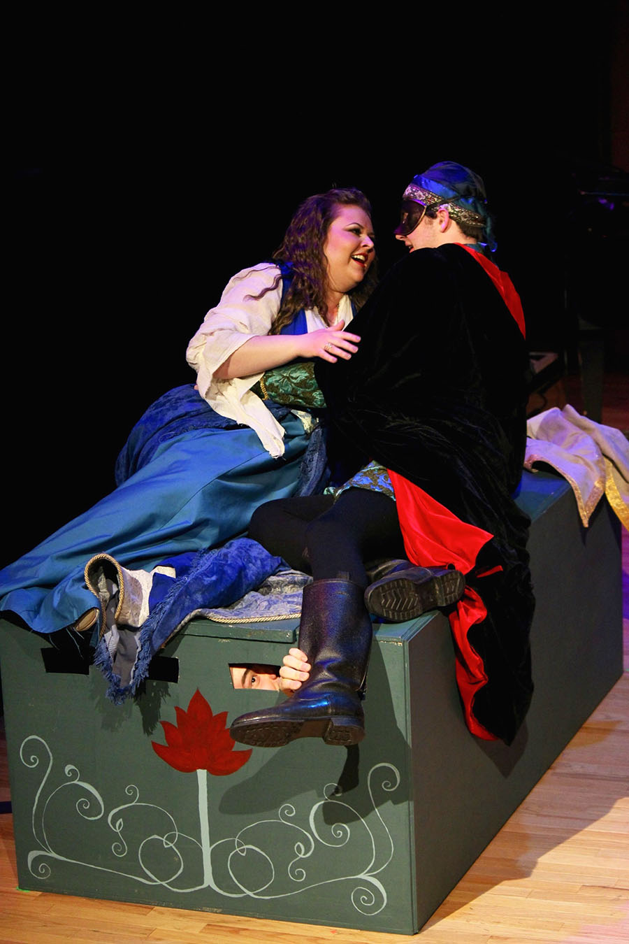 two actors in renaissance costume reclining on bed, smiling. One wears a mask.
