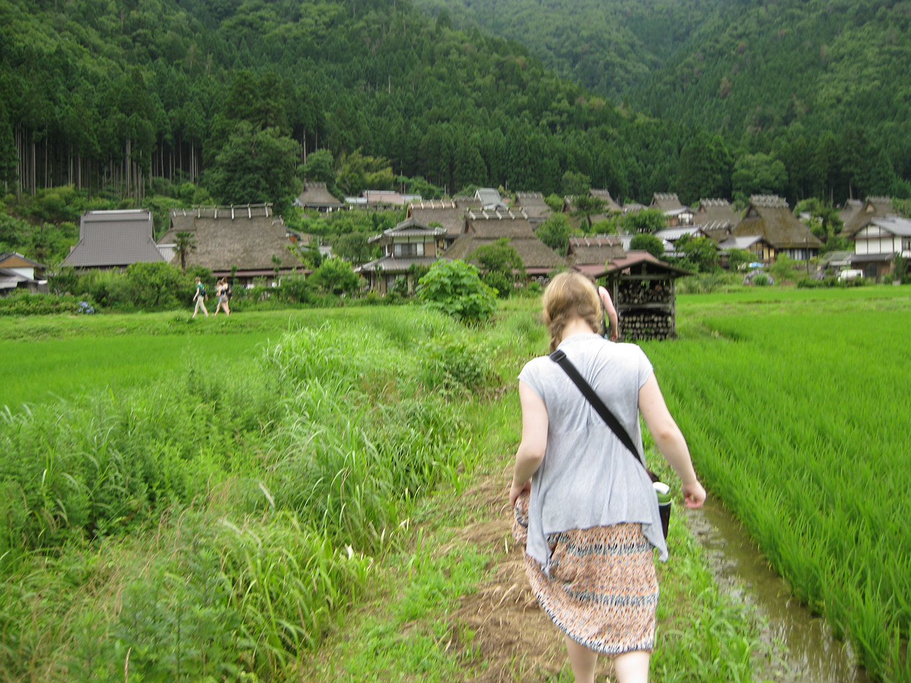 a student walks down a verdant lane toward a rustic village