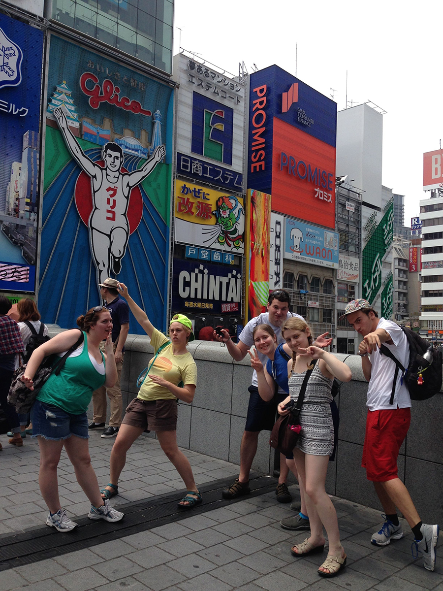 six students strike silly poses in front of Japanese advertisements in an urban setting