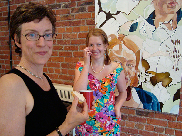 Julia Sapin and student Laura Hudson stand in front of a painting, smiling at the camera. Julia holding a partially eaten bagel and a red cup