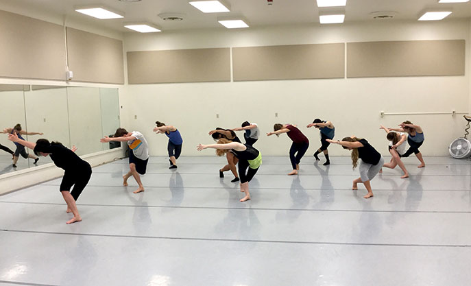 A group of students in a dance classroom doing a bowing pose with one arm and one leg crossed