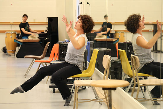 A dance teacher illustrates a pose seated in a chair