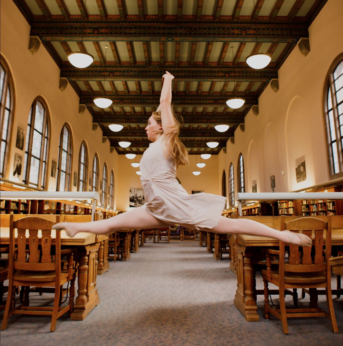 a dancer leaps between tables in an old fashioned library reading room