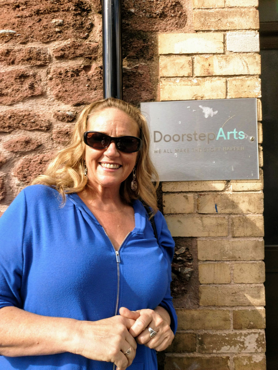 Deb Currier wearing a blue shirt and sunglasses by a entrance with a sign reading Doorstep Arts