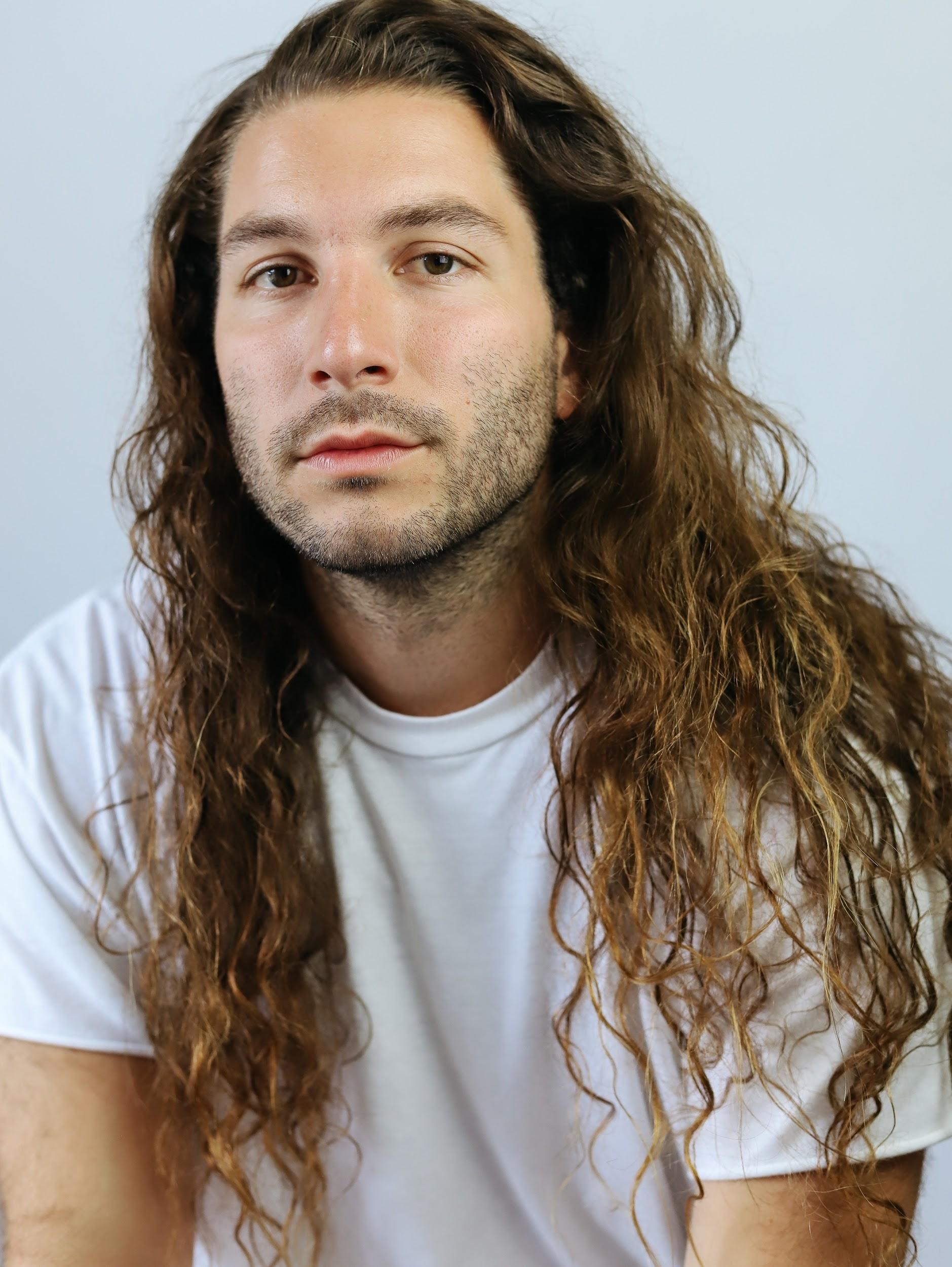young man in a t-shirt with very long, wavy hair and beard and mustache stubble
