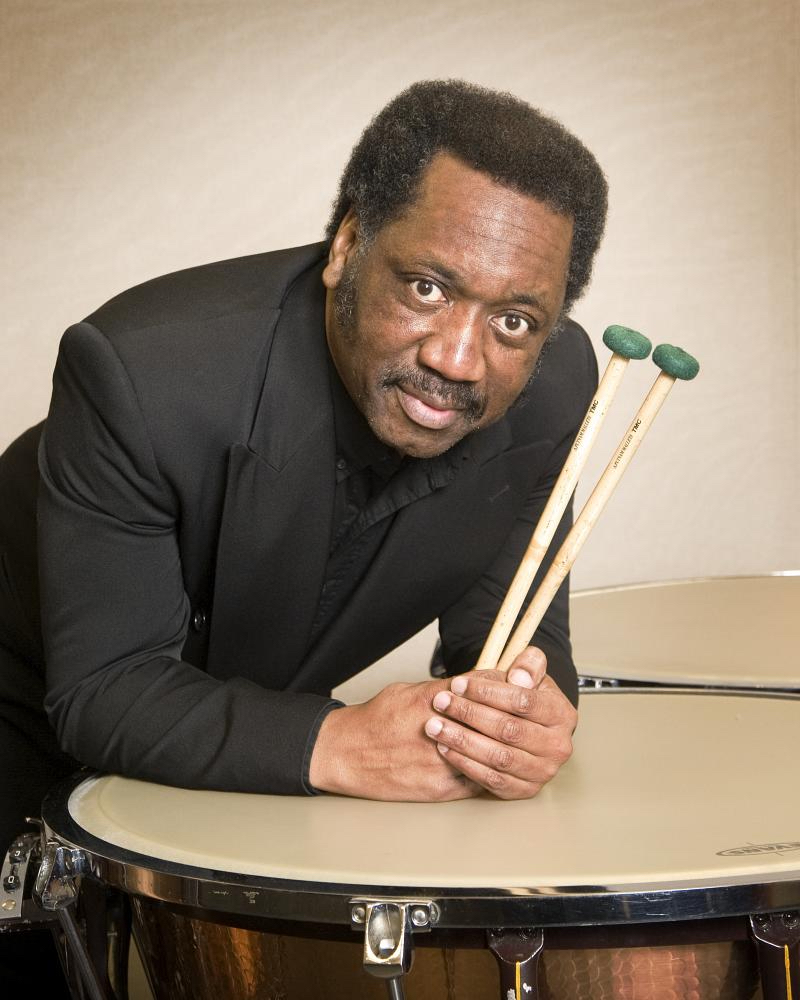 Michael Crusoe in a snazzy formal suit, holding drumsticks in his hands while leaning his elbows on a large drum and looking up at the camera