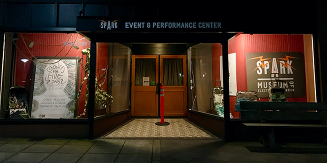 NighLarge display windows with Spark Museum signs and an awning reading "SPARK Event & Performance Center" surround double doors inset from the sidewalk. A traffic cone sits centered in front of the doors.