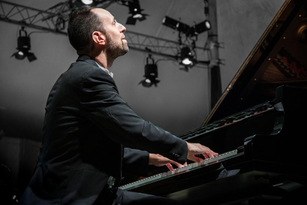 View from the side at floor level: Spencer Myer looks up dreamily as he plays the piano. Stage lights are visible high in the air behind him.