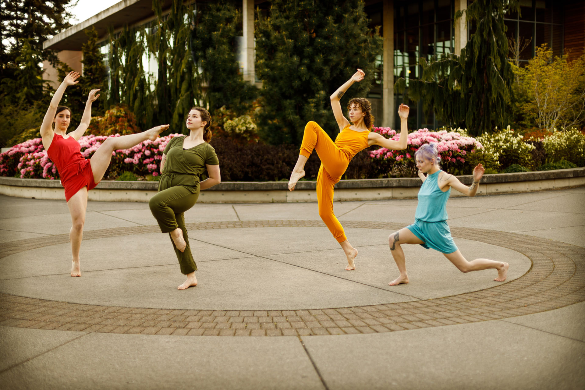 Four dancers posing in different ways inside a circular paved area in front of a garden