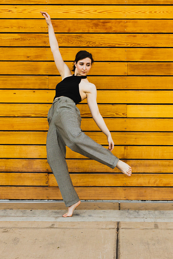 Hannah in posing in front of wood plank wall: leaning back on the ball of one foot with one hand up and the other foot and hand almost touching each other.