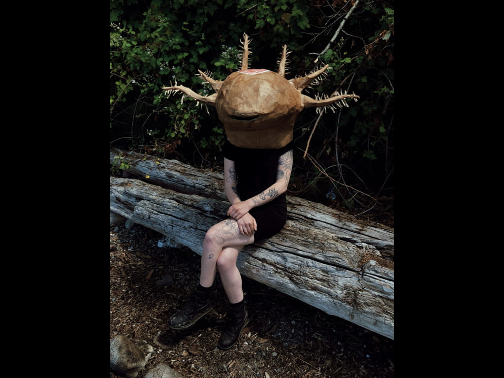 A person sits with crossed legs and hands on a log, wearing a sculpted helmet in the shape of a fantasy creature's head with hairy tentacles