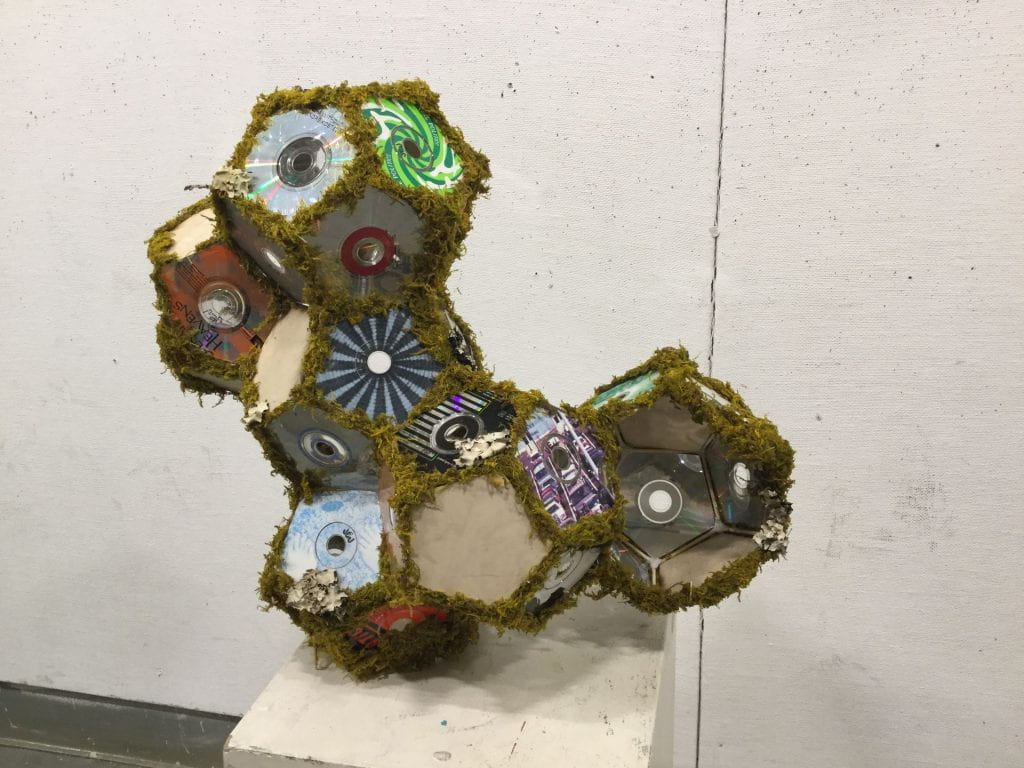 dodecahedrons formed from cardboard and CDs, connected to form a tipped L shape. Each polygonal surface is edged with moss