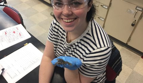 A student wearing safety glasses in a lab classroom holds a frog in a gloved hand and looks up from their desk with an excited smile