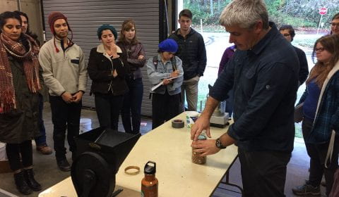 students watching a demonstration in a garage. The presenter is opening a jar of nuts on a table. A grinder is clamped to the end of the table