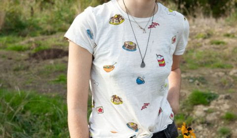 A student smiling at something outside the picture. They are wearing wading boots, a backward baseball cap, a shirt with illustrations of food, and two yellow flowers in their pants pocket.