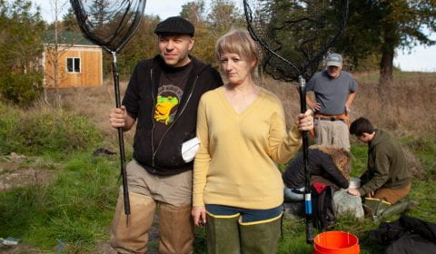 two people in wading boots holding fishing nets in a grassy field. Behind them is a bucket and three people fiddling with something on top of a rock.