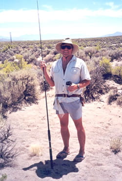  Roger Anderson standing in a desert holding a pole and a coffee mug, wearing a sun hat and sunglasses and smiling