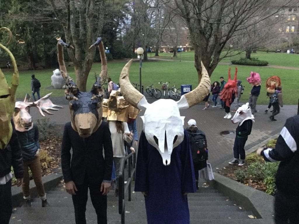 A crowd of people gathered on and around an outdoor staircase, all wearing various animal themed full head masks
