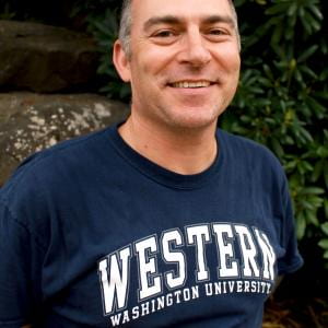 Peter Thut wearing a Western Washington University t-shirt, smiling. green bushes in the background
