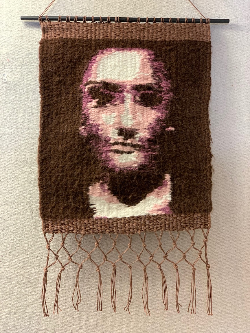 A woven tapestry hangs from a dowel. The tapestry's pattern creates an image of a woman's face