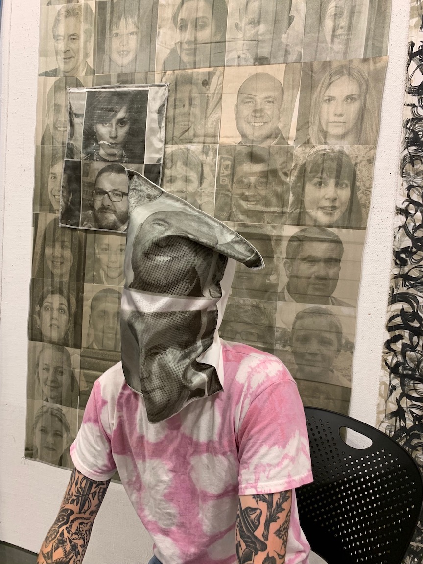 A person in a tie-died t-shirt and tatoo-covered arms sits with a bag over their head which has faces printed on it. On the wall behind hangs a collage of several printed faces.