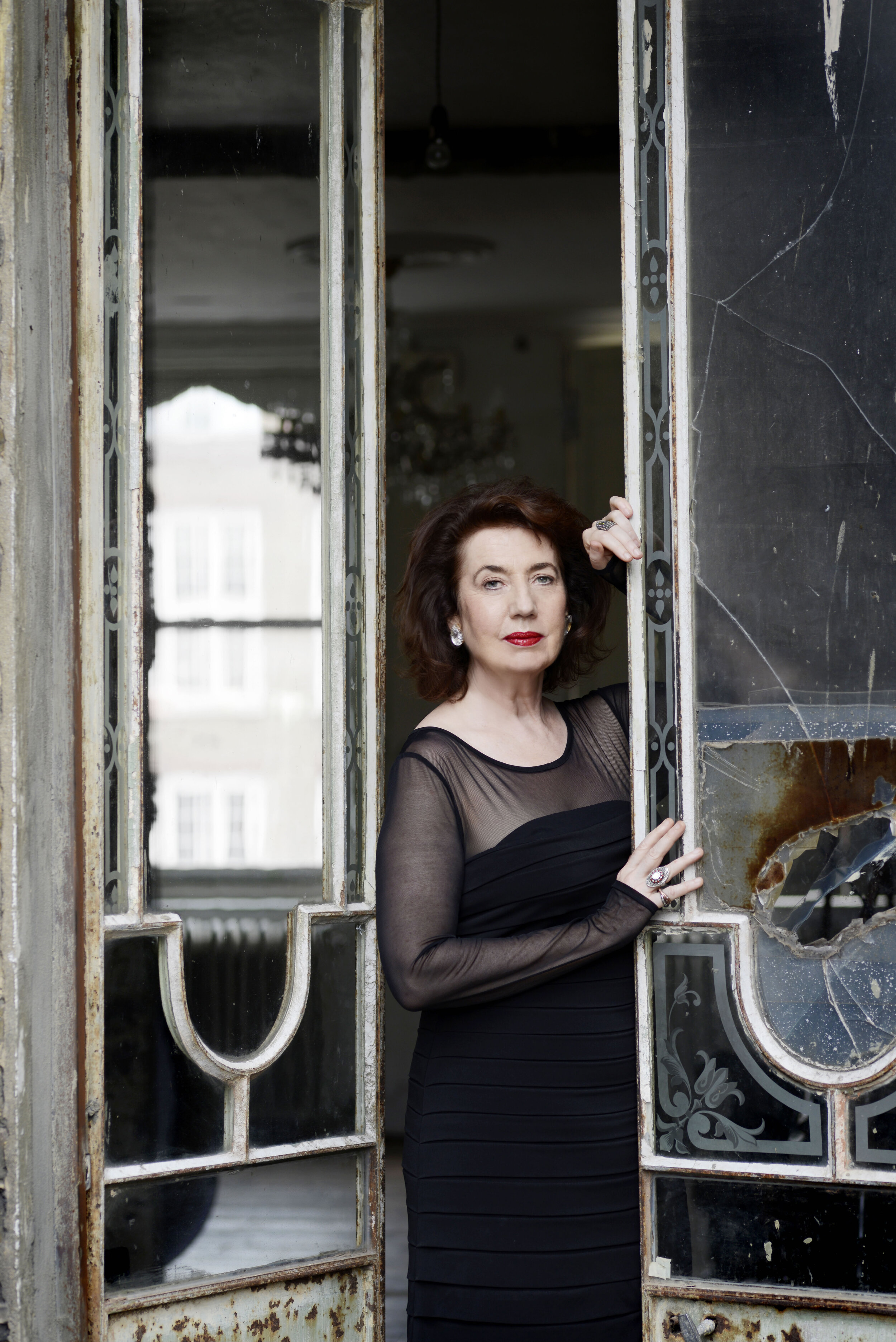  Imogen Cooper in formal attire with a calm, thoughtful face, standing in the opening of an antique double glass door (missing some glass) of an abandoned building, lightly leaning on one door.