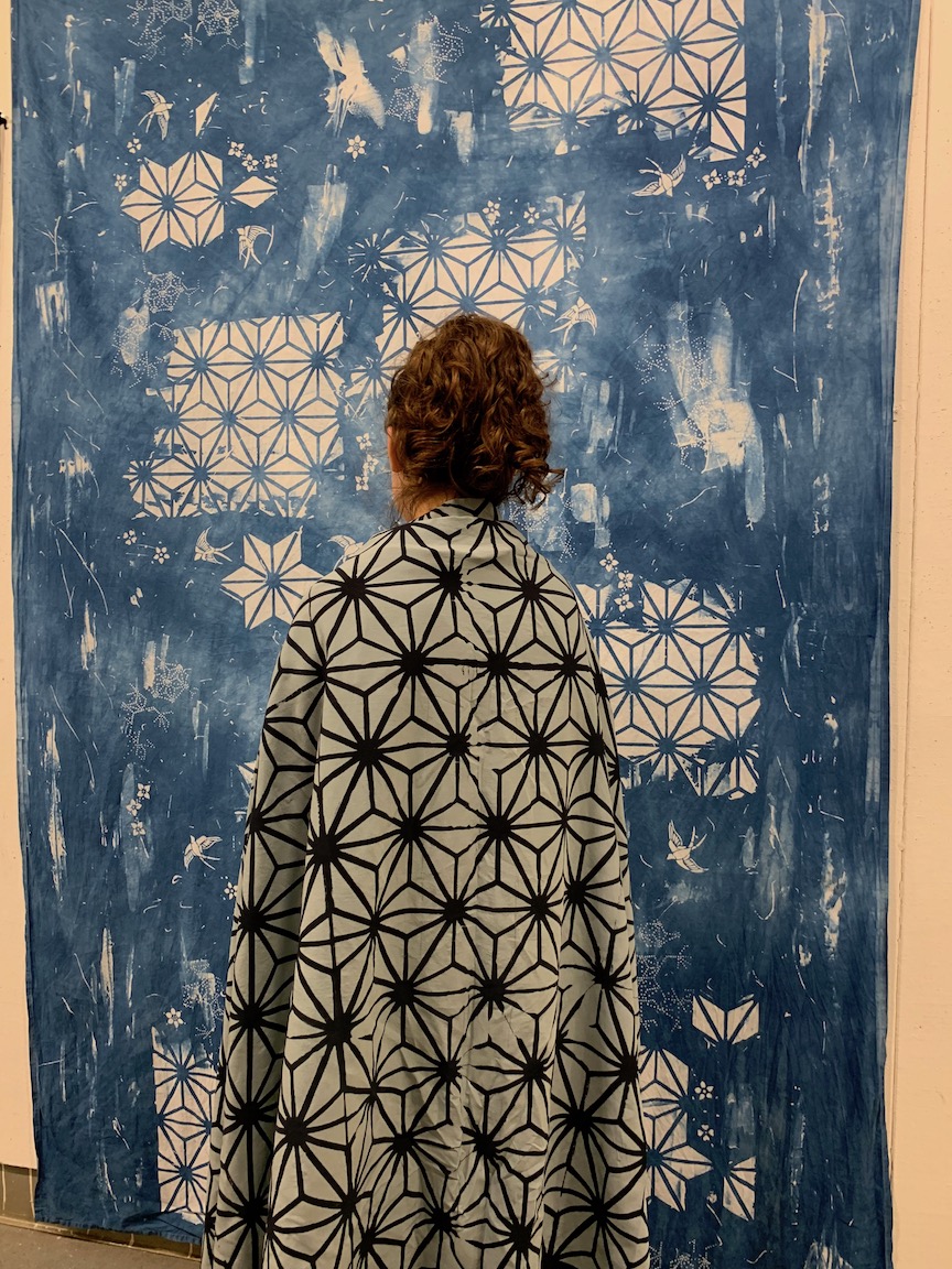 Backside of a person wearing a blanket with geometric print, standing in front of a large piece of cloth hanging on a wall with similar printing