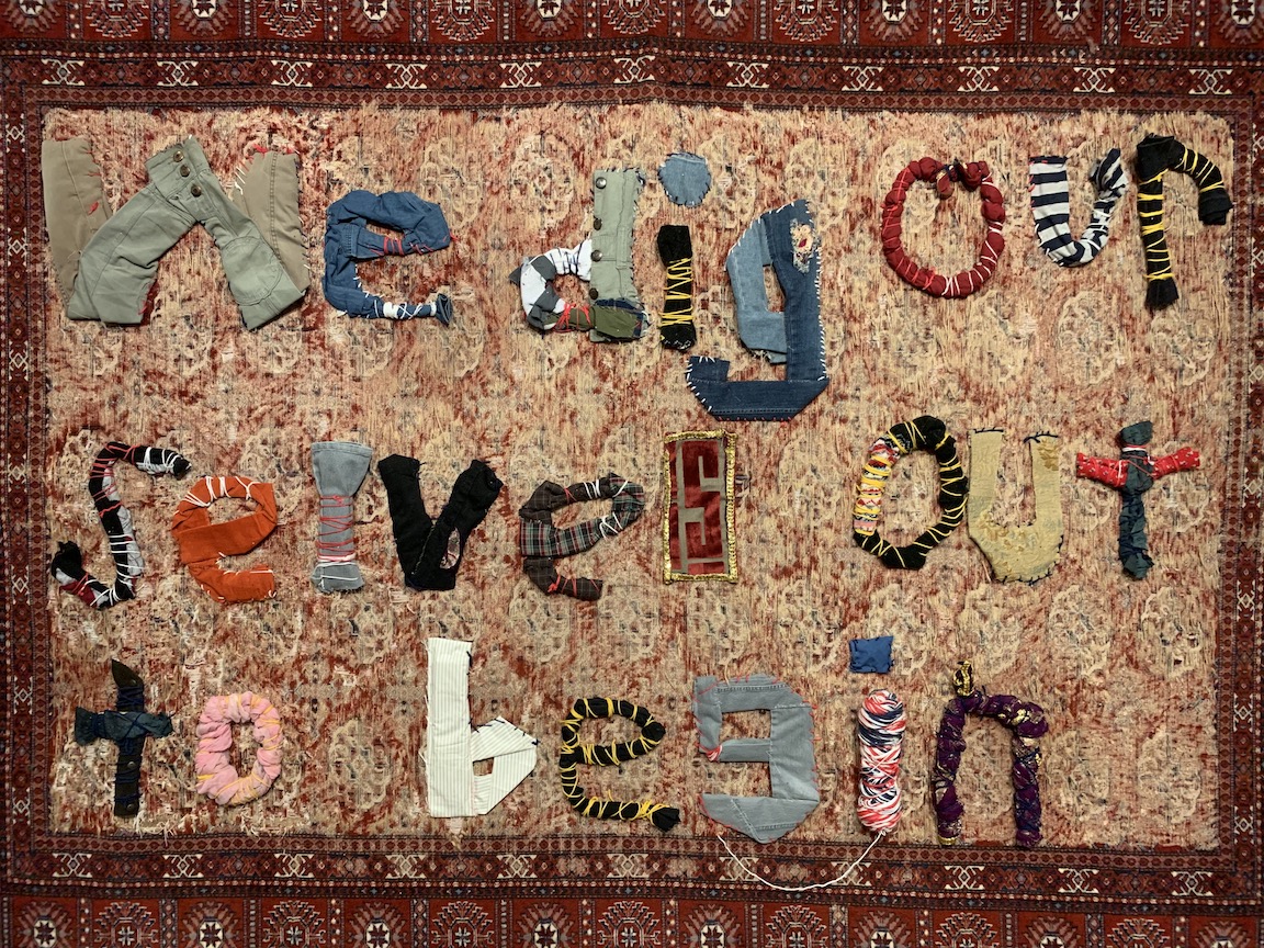 A quilt or tapestry with letters sewn on to it, spelling out a message: "we dig ourselves out to begin"