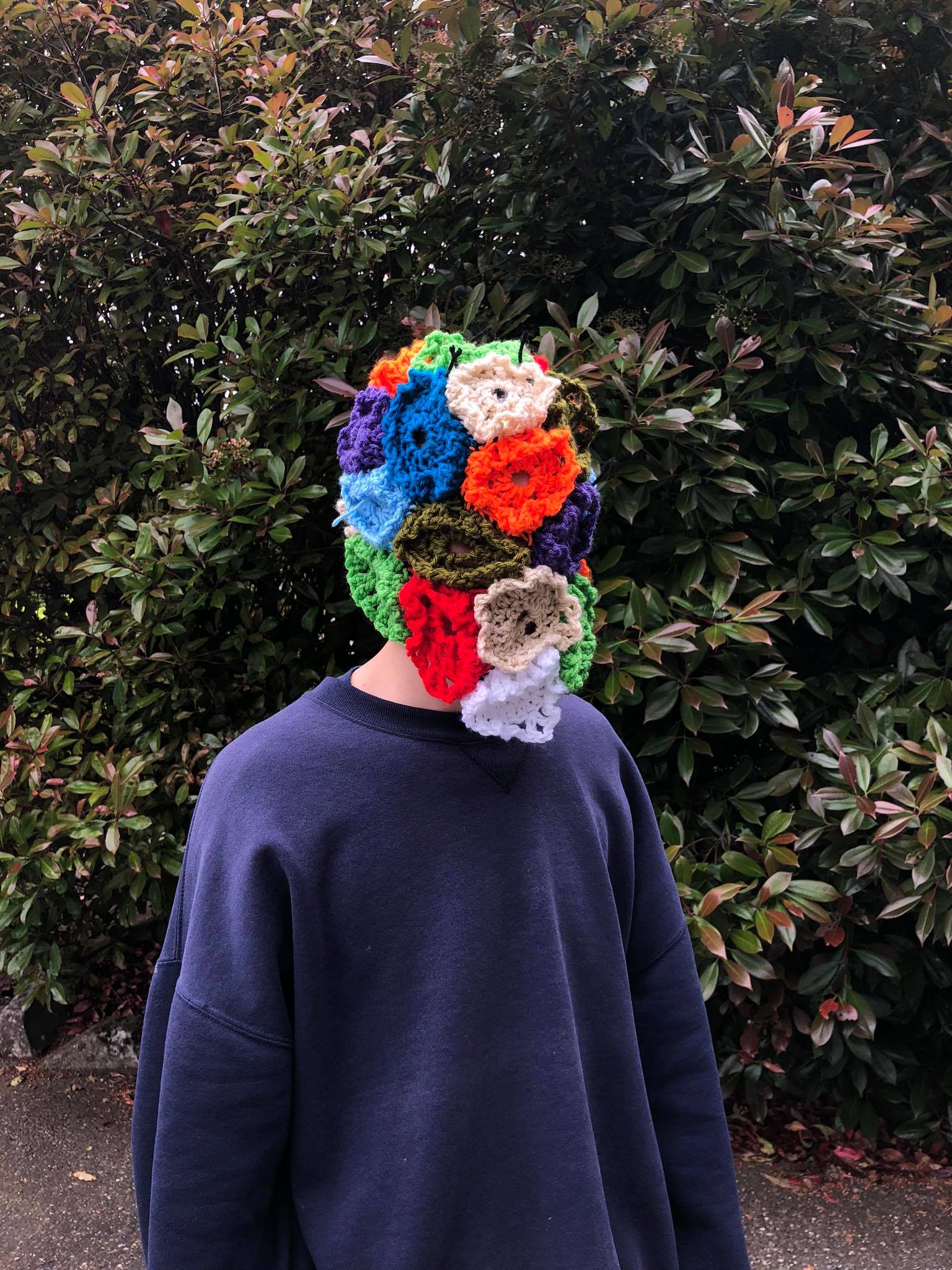 A person standing outside wearing a face-covering helmet made of knitted doilies
