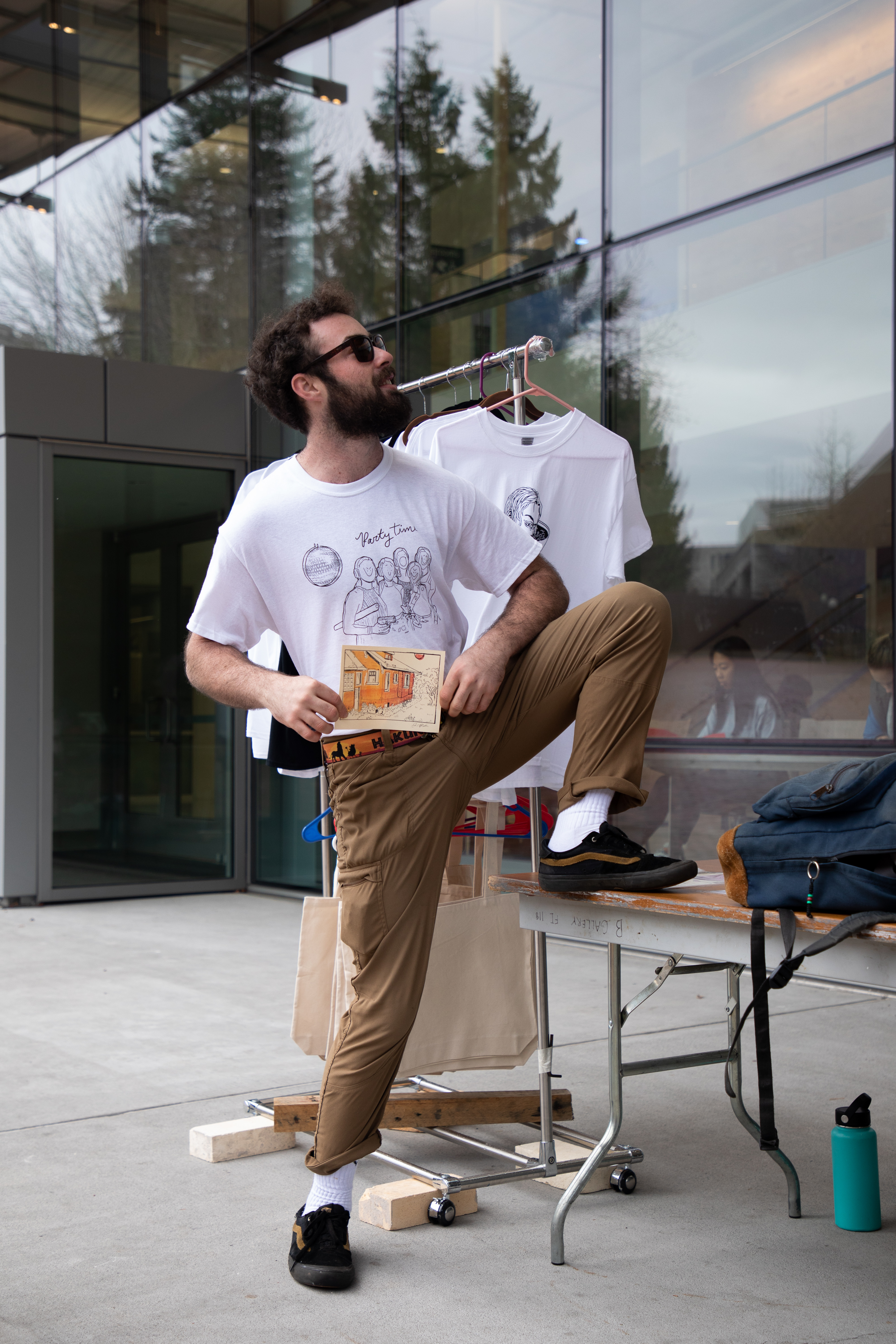 A person wearing sunglasses and a screenprinted shirt, standing next to an outdoor display table with one foot on the table, proudly holding a small drawing and looking up and to the side with a smile. A rack of screenprinted t-shirts and tote bags hangs behind the person, next to the table.