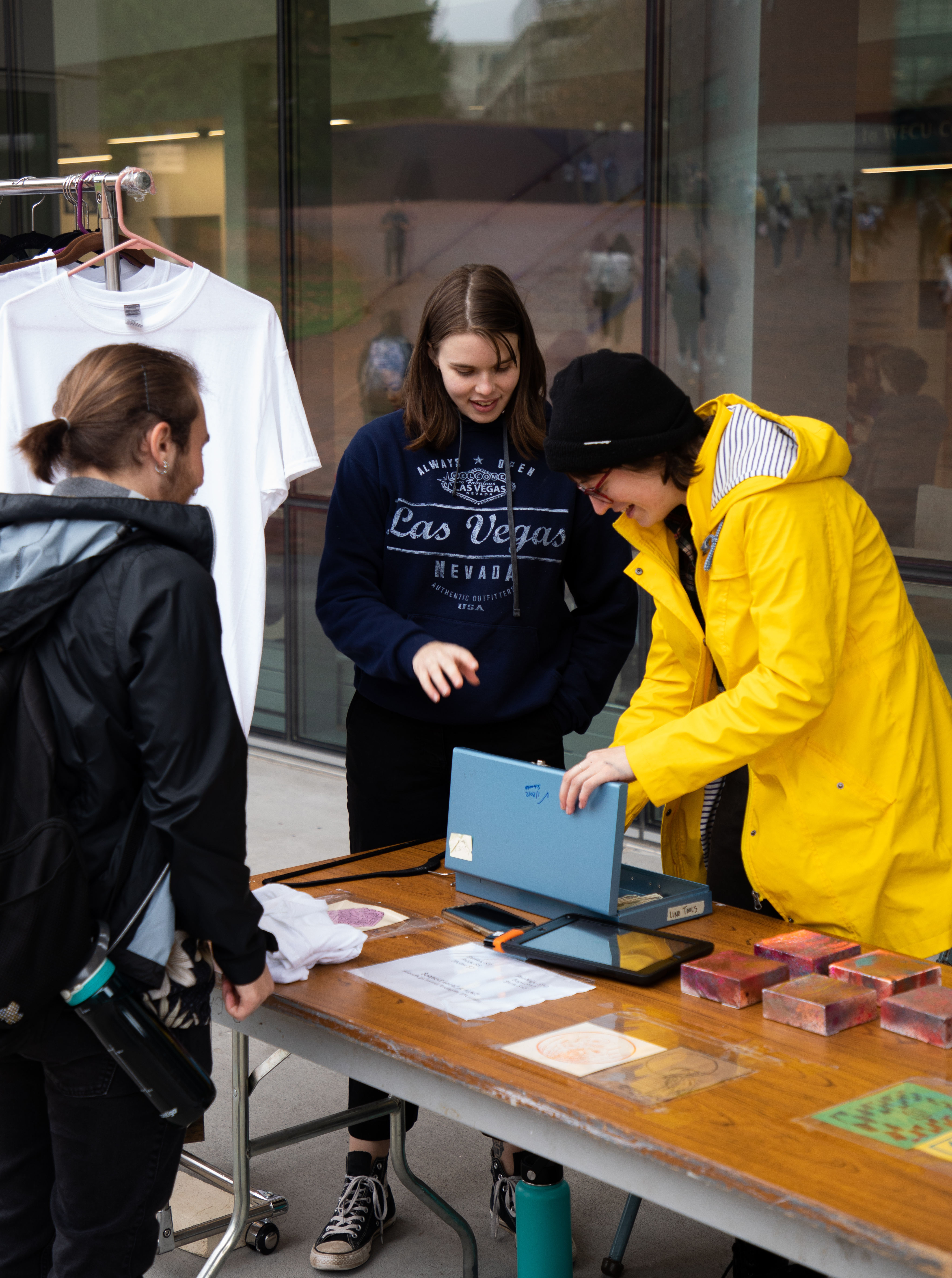A person waits with a folded shirt at a display table while two people examine the contents of a cash box from the other side of the table. All are smiling.