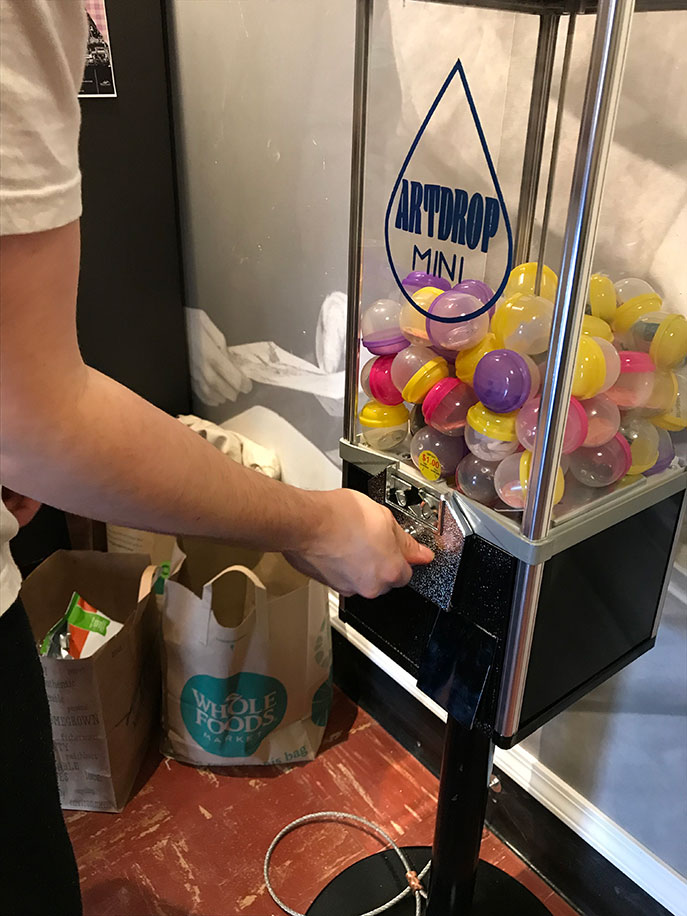a hand turns the crank on a small art vending machine filled with colorful plastic eggs of art