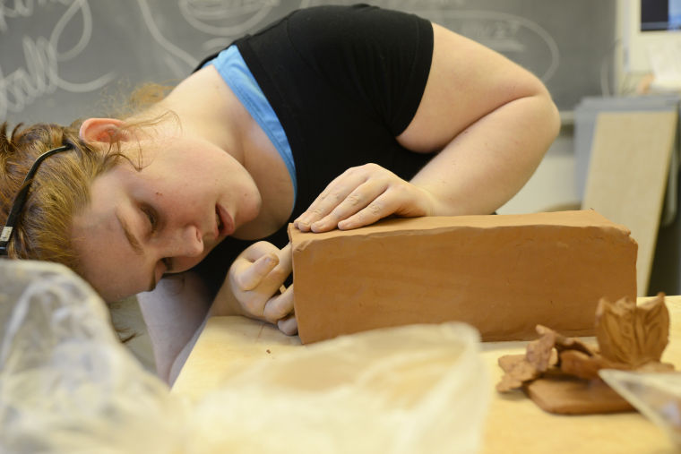 A student leans to the side with their face up close to the end of a block of clay which is the size of a loaf of bread