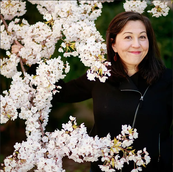 a smiling poet in a black top appears from behind a treeful of pale blossoms