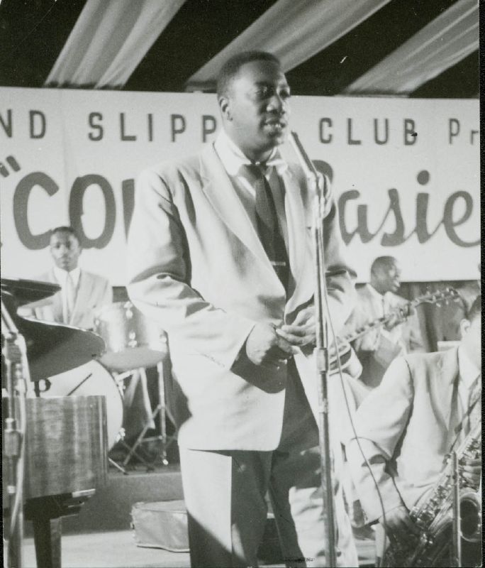 Count Basie in a suit and tie, stands on stage with his hands together, vocalizing into a microphone. Behind him, band members are busy playing saxophone, guitar and drums. Above the band is a sign spanning the stage and almost as tall as a person which reads "Shoe 'n' Slipper Club: "Count Basie""